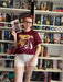 a woman in a red shirt and white shorts standing in front of a book shelf